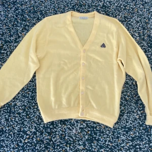 Gul kofta från IZOD - Säljer en ljusgul kofta från IZOD med knappar framtill och en broderad logga på bröstet. 