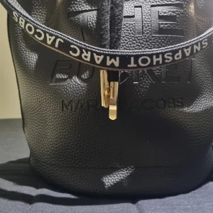 Svart handväska från Marc Jacobs - Snygg svart handväska från Marc Jacobs med textdetaljer och guldfärgad dragkedja. Väskan har en justerbar axelrem med logotyp och en stilren design som passar perfekt för alla tillfällen.