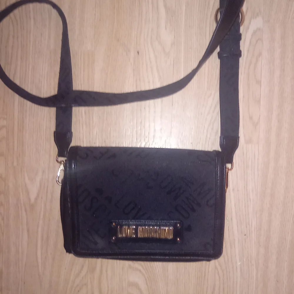 Snygg svart axelväska från Love Moschino med ett subtilt mönster och gulddetaljer. Väskan har en justerbar axelrem och ett rött innerfoder som ger en lyxig känsla. Perfekt för att bära dina nödvändigheter med stil. Nypris ca 900 kr. Laukut & Käsilaukut.