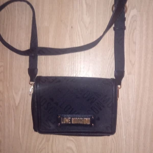 Svart axelväska från Love Moschino - Snygg svart axelväska från Love Moschino med ett subtilt mönster och gulddetaljer. Väskan har en justerbar axelrem och ett rött innerfoder som ger en lyxig känsla. Perfekt för att bära dina nödvändigheter med stil. Nypris ca 900 kr
