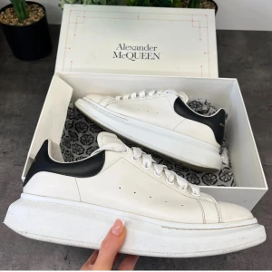 Vita sneakers från Alexander McQueen - Snygga vita sneakers från Alexander McQueen med svart häl och märkets logga i guld. Skorna har en chunky sula och klassisk snörning. Perfekta för en stilren look.