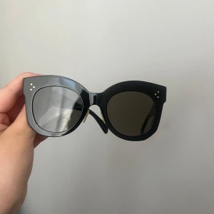 Svarta solglasögon från Céline - Snygga svarta solglasögon från Céline med en elegant och stilren design. De har en bred båge och små guldfärgade detaljer vid hörnen. Perfekta för att ge en lyxig touch till din outfit.