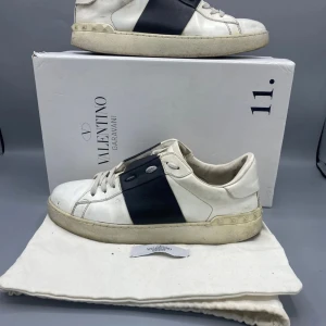 Valentino opens - Snygga vita sneakers med en bred svart rem över mitten. Skorna har en klassisk design med låg profil och är perfekta för en stilren look. Får med nya skosnören
