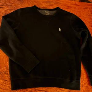 Svart tröja från Ralph Lauren - Säljer en stilren svart tröja från Ralph Lauren med det klassiska logotypbroderiet på bröstet. Tröjan har långa ärmar och en rund halsringning. Perfekt för en avslappnad och snygg look.