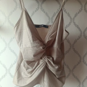 Beige topp med knut från BikBok - Säljer en beige ribbad linnetopp från BikBok med smala justerbara axelband och snygg knutdetalj framtill. Toppen har en lätt glansig yta och är perfekt för varma dagar i sommar. Axelband som går att ändra. Använd ett fåtal gånger, 40 kr