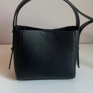 Svart axelväska i skinnimitation - Snygg svart axelväska med stilren design och fyrkantig form. Väskan har ett långt axelband och stängs med magnetknapp. Perfekt storlek för det viktigaste och passar till många olika outfits.