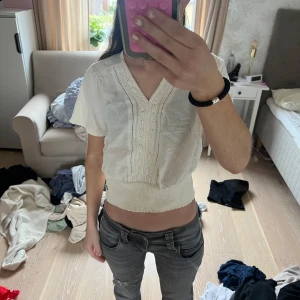 Vit croppad blus med spetsdetaljer - Säljer en söt vit croppad blus med v-ringning och spetsdetaljer framtill. Blusen har korta ärmar och bred smockad midja som ger en fin passform. Perfekt för vår och sommar!