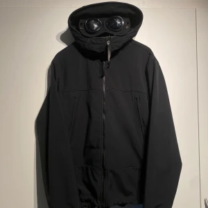 C.P. Company Shell-R Goggles Jacket  - Säljer en svart jacka från C.P. Company med ikoniska goggles i huvan. Jackan har dragkedja framtill, två sidofickor och en normal passform.   Skal material, vatten och vindtät. Storlek 56 vilket motsvarar XL/L. Nypris 5000kr. Välanvänd men i okej skick. Fler bilder finns i DM! 