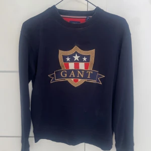 Mörkblå sweatshirt från GANT - Säljer en mörkblå sweatshirt från GANT med stort broderat sköldmärke och text på bröstet. Tröjan har rund halsringning och långa ärmar. Klassisk och stilren design med ribbade muddar.