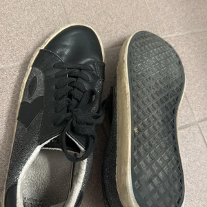 Svarta sneakers från Le Temps Des Cerises med glitterdetaljer - Säljer ett par svarta sneakers från Le Temps Des Cerises med glittriga sidor och svarta snören. Skorna har vit sula och svart baksida. Säljer på grund av att det är för små!!