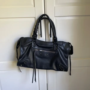 Svart handväska  - Säljer en svart handväska i skinnimitation med snygga flätade handtag och dragkedja upptill. Väskan har dekorativa nitar och långa remmar framtill samt innerfack för småsaker. Små defekter, se bild 4 och 5. Priset kan förhandlas, skriv vid frågor.💞