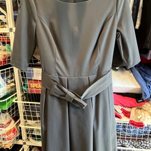 Mörkgrå klänning med rosett från Sisley - Stilren mörkgrå klänning från Sisley med knälång passform, trekvartsärm och markerad midja med brett bälte och dekorativ rosett framtill. Klänningen har rund halsringning och är tillverkad i ett mjukt, slätt material.