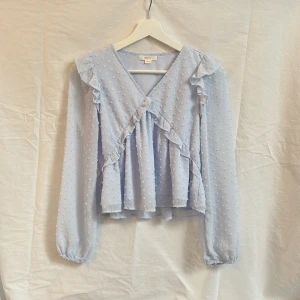 Ljusblå blus med volang och prickigt mönster - Supersöt ljusblå blus med volangdetaljer och diskret prickigt mönster. Blusen har v-ringning, långa ärmar med resår vid handlederna och en luftig passform. Perfekt för en romantisk look!