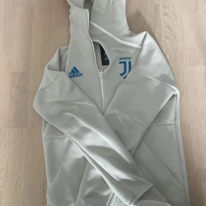 Ljusgrå hoodie från Adidas x Juventus - Säljer en ljusgrå hoodie från Adidas i samarbete med Juventus. Jackan har dragkedja, huva och blå loggor på bröstet. Perfekt för dig som gillar fotboll och vill ha en stilren look.