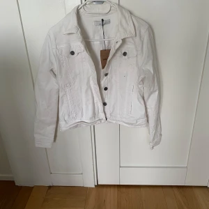 Vit jeansjacka med knappar - Säljer en klassisk vit jeansjacka med silverfärgade knappar framtill. Jackan har två bröstfickor med lock och knapp. Perfekt att slänga över en t-shirt eller klänning för en fräsch look.
