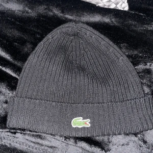 Svart ribbad mössa från Lacoste - Svart ribbstickad mössa från Lacoste med klassisk grön krokodillogga framtill. Tillverkad i 100% ull, enkel och stilren design som passar till det mesta. Perfekt för kyliga dagar.