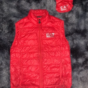 Röd dunväst från Emporio Armani EA7 - Snygg röd dunväst från Emporio Armani EA7 med silverfärgad logga på bröstet. Västen har hög krage och dragkedja framtill. Väldigt bra för sommaren. Det med kommer en dust bäg.