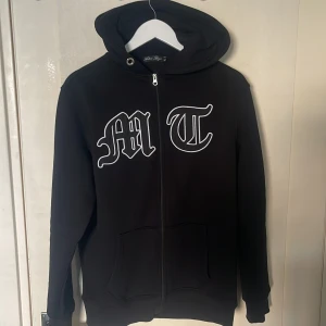 TUTTOLENTE HOODIE - Säljer en svart hoodie från Tuttolente med vita bokstäver 'MT' på bröstet och en stor svart örn på ryggen. Tröjan har dragkedja framtill, huva och två fickor. Perfekt för en avslappnad stil.