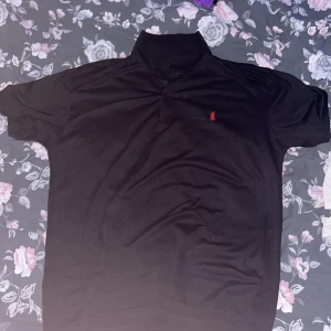 Svart pikétröja från Polo Ralph Lauren - Säljer en klassisk svart pikétröja från Polo Ralph Lauren med röd broderad logga på bröstet. Tröjan har korta ärmar, krage och knappar vid halsen. Perfekt för en stilren och avslapnad stil 
