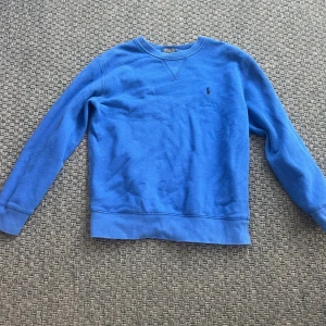 Blå sweatshirt från Polo Ralph Lauren - Säljer en klassisk blå sweatshirt från Polo Ralph Lauren med broderad logga på bröstet. Tröjan har rund halsringning och långa ärmar, perfekt för en avslappnad stil.
