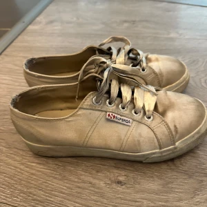 Beige sneakers från Superga - Säljer ett par klassiska beige sneakers från Superga med snörning och rund tå. Skorna har en platt sula och är tillverkade i canvasmaterial. Perfekta för en avslappnad stil och passar till det mesta.