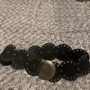 Svart bälte med stora runda nitar och silverfärgat spänne - Unikt svart bälte med stora runda läderbitar och dekorativa silverfärgade nitar. Bältet har ett stort, rundat spänne med ingraverat mönster. Perfekt accessoar för att lyfta din outfit.