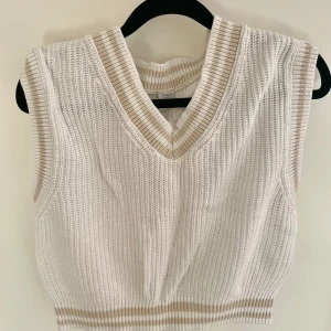 Vit stickad väst med beigea detaljer från Lindex - Vit stickad väst i storlek 134/140 från Lindex. Västen har ribbade detaljer i beige vid halsringning, ärmöppningar och nederkant. V-ringad och ärmlös, perfekt att ha över en långärmad tröja eller skjorta! 