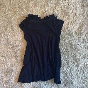 Mörkblå topp med volangkrage från Holly & White - Säljer en mörkblå topp från Holly & White med volangdetaljer runt kragen. Toppen är ärmlös och har en elegant, enkel design som passar perfekt till sommaren. Materialet känns mjukt och lätt, och modellen är normal i passformen.