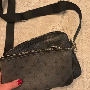 Svart axelväska från Louis Vuitton - Säljer en svart axelväska från Louis Vuitton med klassiskt monogrammönster. Väskan har justerbar axelrem, två fack med dragkedja och en liten avtagbar plånbok. Perfekt för att bära med sig det viktigaste. Äkta men ganska sliten nypris 22 tusen men säljer för 5000