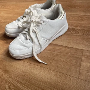 Vita sneakers med gulddetalj - Säljer ett par fräscha vita sneakers med snygg gulddetalj vid hälen. Perfekta för dig som vill ha en stilren och trendig look till vardags.