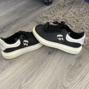 Svarta och vita sneakers med Karl Lagerfeld-tryck - Säljer ett par svarta och vita sneakers från Karl Lagerfeld med ikoniskt tryck på sidan. Skorna har rund tå, platt sula och svart snörning. Perfekta för dig som vill sticka ut med en cool detalj på fötterna.