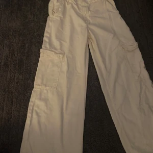 Beiga cargopants från H&M - Säljer ett par beiga cargopants från H&M med bred passform och stora fickor på sidorna. Byxorna har resår i midjan och är perfekta för en avslappnad stil. Klassisk cargomodell i mjukt bomullsmaterial.