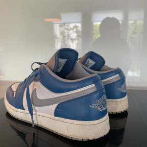 Nike Air Jordan 1 Low blå och vita sneakers - Säljer ett par Nike Air Jordan 1 Low sneakers i blått, vitt och grått. Skorna har klassisk snörning,platt sula. Perfekta för dig som gillar streetwear och vill ha en ikonisk sneaker i garderoben.