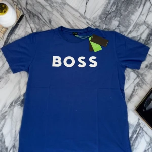 🎉💙🔥Helt Ny! Blå t-shirt från BOSS - Snygg blå t-shirt från BOSS med vit logotyp tryckt framtill. Klassisk rund halsringning och normal passform. Perfekt för en stilren och enkel look.