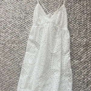 Vit spetsklänning med smala axelband från H&M - Superfin vit klänning från H&M med smala axelband och v-ringning. Klänningen har broderade spetsdetaljer och ett vackert hålmönster över hela tyget. Perfekt för sommarens alla tillfällen!
