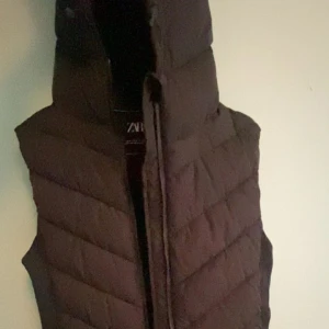 Svart väst från ZARA - Säljer en svart dunväst från ZARA med huva. Västen har ett quiltat mönster och stängs med dragkedja framtill. Perfekt att slänga över en hoodie eller tröja för extra värme.