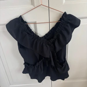 Svart volangtopp med rosett - Säljer en svart topp med bred volang runt axlarna och en stor rosett framtill. Toppen har en luftig passform och är perfekt för att lyfta en outfit. Materialet känns lätt och svalt.