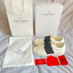 Valentino open - Säljer ett par vita Valentino Garavani sneakers med bred svart rem över vristen och vita snören. Skorna har rund tå, platt sula och ikoniska nitar på hälen. Kommer med originalkartong, dustbag och certifikat. Storlek 42, men kan även passa 41 eller 43. Skriv vid några mer frågor eller funderingar. OBS; 1nte 0rg1nal‼️