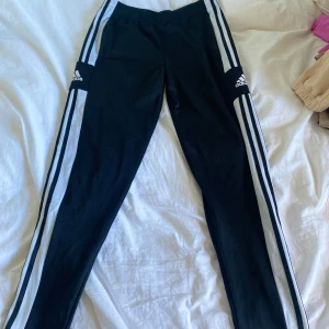Svarta Adidas träningsbyxor med vita ränder - Säljer ett par klassiska svarta Adidas träningsbyxor med vita ränder längs sidorna och Adidas-logga på båda benen. Byxorna har en sportig look och är perfekta för träning eller chill. Resår i midjan för skön passform.