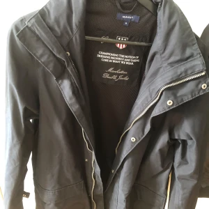 Mörkblå vindjacka från Gant - Säljer en svart vindjacka från Gant med huva och dragkedja. Jackan har stora fickor framtill och detaljer i form av knappar och dragkedja. Perfekt för blåsiga dagar och enkel att matcha med olika outfits.