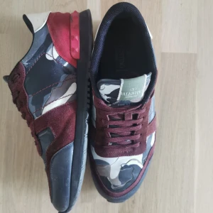 Valentino Rockrunner  - Säljer ett par Valentino Rockrunner sneakers med snygga röda och vinröda detaljer samt camouflage-mönster i blått, grått och vitt. Skorna har snörning och en platt sula. Perfekta för dig som vill sticka ut med stil.