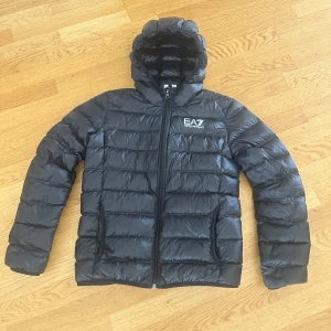 Svart dunjacka med huva från EA7 - Säljer en svart dunjacka från EA7 med huva och dragkedja framtill. Jackan har ett stilrent, quiltat mönster och EA7-logga på bröstet. Perfekt för kyligare dagar och enkel att matcha med olika outfits.