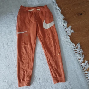 Orange mjukisbyxor från Nike - Säljer ett par orange mjukisbyxor från Nike med stora vita swoosh-loggor på både fram- och baksida. Byxorna har resår i midjan och muddar vid bensluten. Perfekta för en avslappnad stil och har tryckdetaljer längs benet.