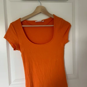 Orange ribbad topp med kort ärm - Säljer en snygg orange ribbad topp med korta ärmar och djup rundad urringning. Perfekt för att liva upp garderoben med en färgklick! Toppen har en figurnära passform och är superbekväm.