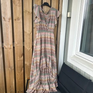 Mönstrad långklänning med volangärmar - Säljer en långklänning med färgglatt sicksackmönster i rosa, grön, gul och beige. Klänningen har volangdetaljer vid ärmarna och v-ringning framtill. Resår i midjan för en smickrande passform och volangkant nertill. Kortare fram. Passar mer Medium men står m/L