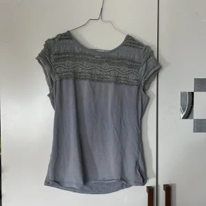 Ljusgrå topp med spetsdetaljer - Säljer en ljusgrå topp med vackra spetsdetaljer upptill och korta ärmar. Toppen har rund halsringning och är tillverkad i ett mjukt material som känns skönt mot huden. Perfekt för dig som gillar romantiska detaljer och en stilren look.