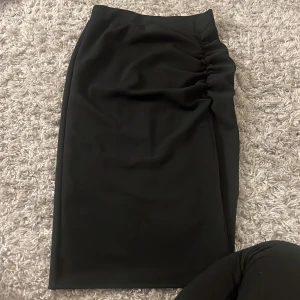 Svart kjol med rynkade detaljer från SHEIN - Svart kjol från SHEIN med snygg rynkning på ena sidan. Kjolen har en rak passform och är knälång, perfekt för en stilren look. Tillverkad i ett mjukt syntetmaterial som känns bekvämt hela dagen.