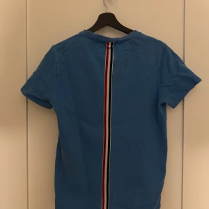 Blå t-shirt från Thom Browne - Säljer en blå t-shirt från Thom Browne med en karakteristisk röd, vit och blå rand som går längs ryggen. Klassisk rund halsringning och korta ärmar. Tillverkad i Italien.