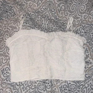 Vit croppad linnetopp med smock - Säljer en söt vit croppad linnetopp med smock och tunna axelband. Toppen har volangdetalj upptill.