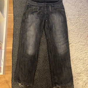 Svarta jeansbyxor med tvättad look - Säljer ett par svarta jeansbyxor med tvättad och något sliten look. Klassisk femficksmodell med dragkedjegylf och knapp i midjan. Passar dig som gillar en lite mer avslappnad stil. Passar om man e 2000s fashion.:)